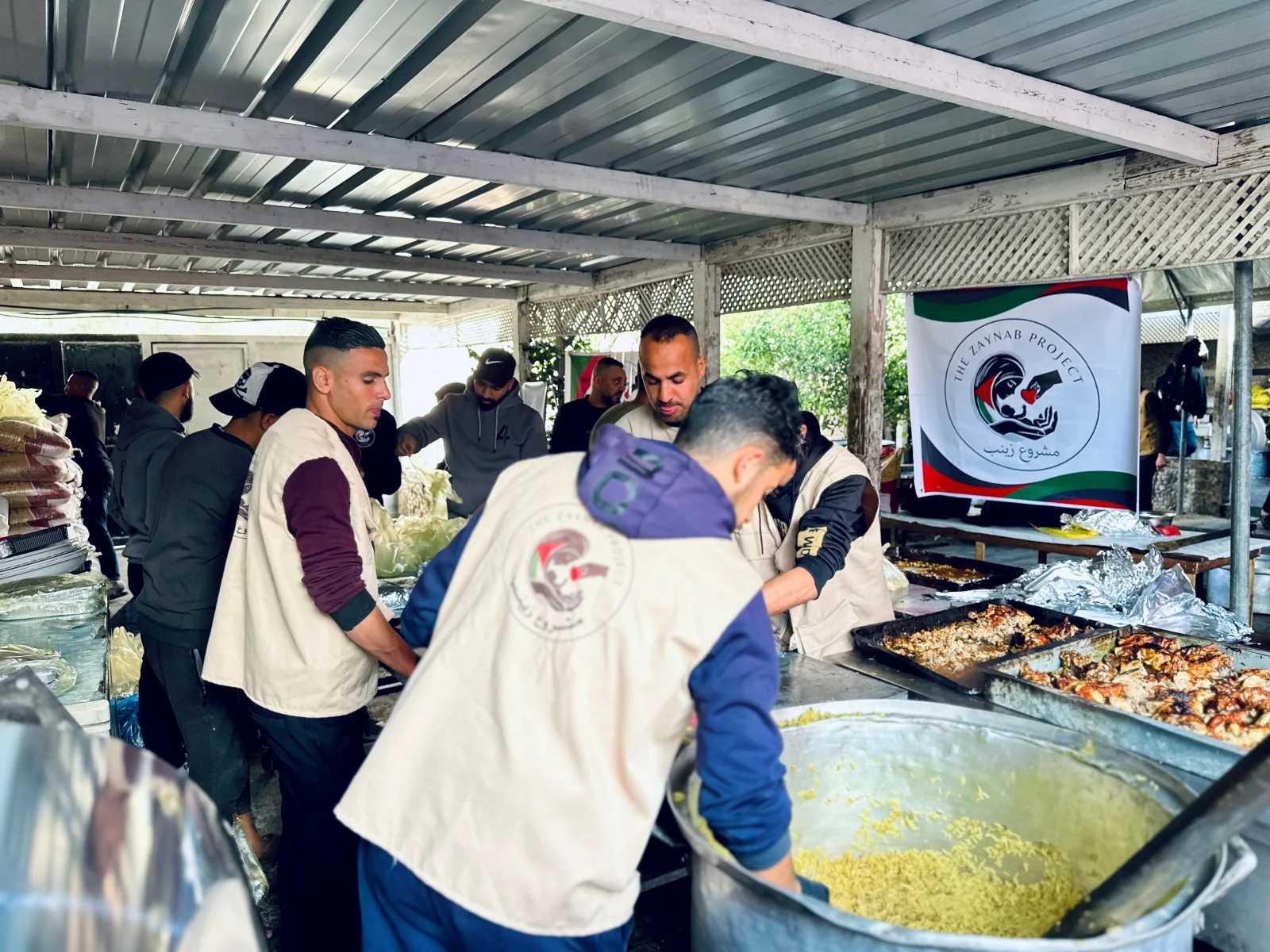 Zaynab Project team members prepare rice and chicken in large cooking vessels beneath a covered kitchen area.
