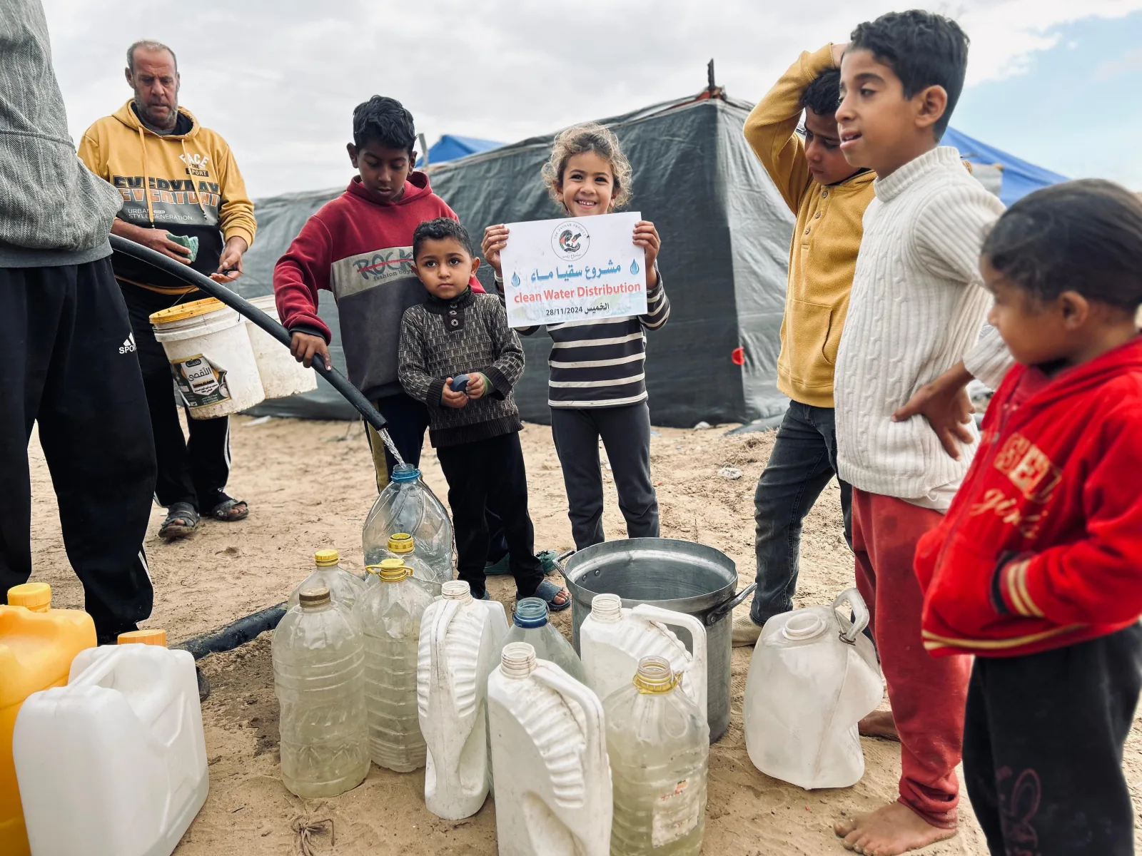 An adult fills water containers while children gather beside tents during a Zaynab Project clean-water distribution.